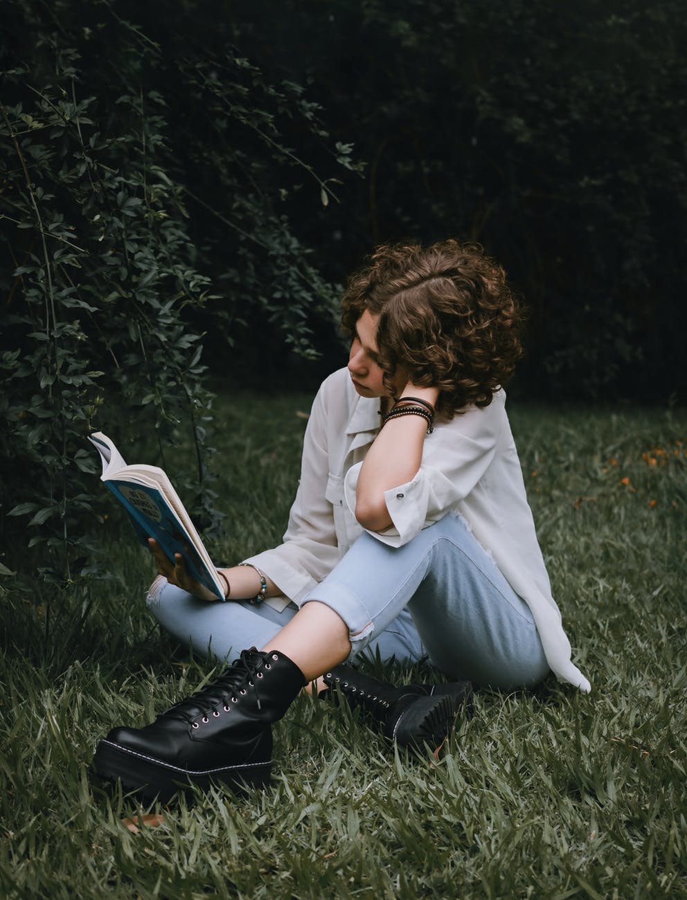 lady reading book while sitting on grassy lawn near plants