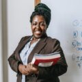 happy black female teacher standing with workbooks near whiteboard